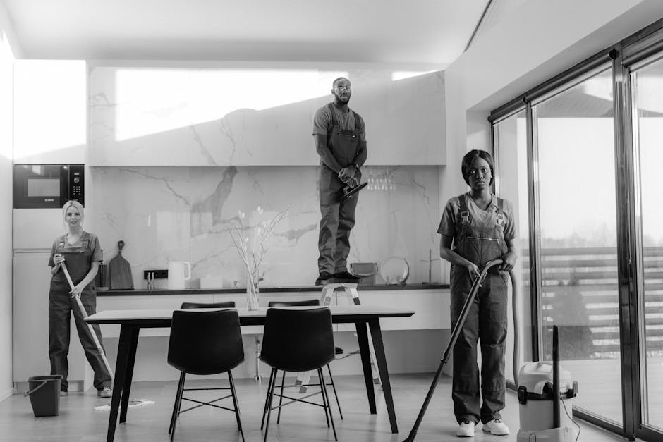 A female domestic cleaner wearing a yellow uniform, blue gloves, and a lanyard with an ID badge, holding a spray bottle and a blue cleaning cloth. She is standing in a modern living room with a sofa, 